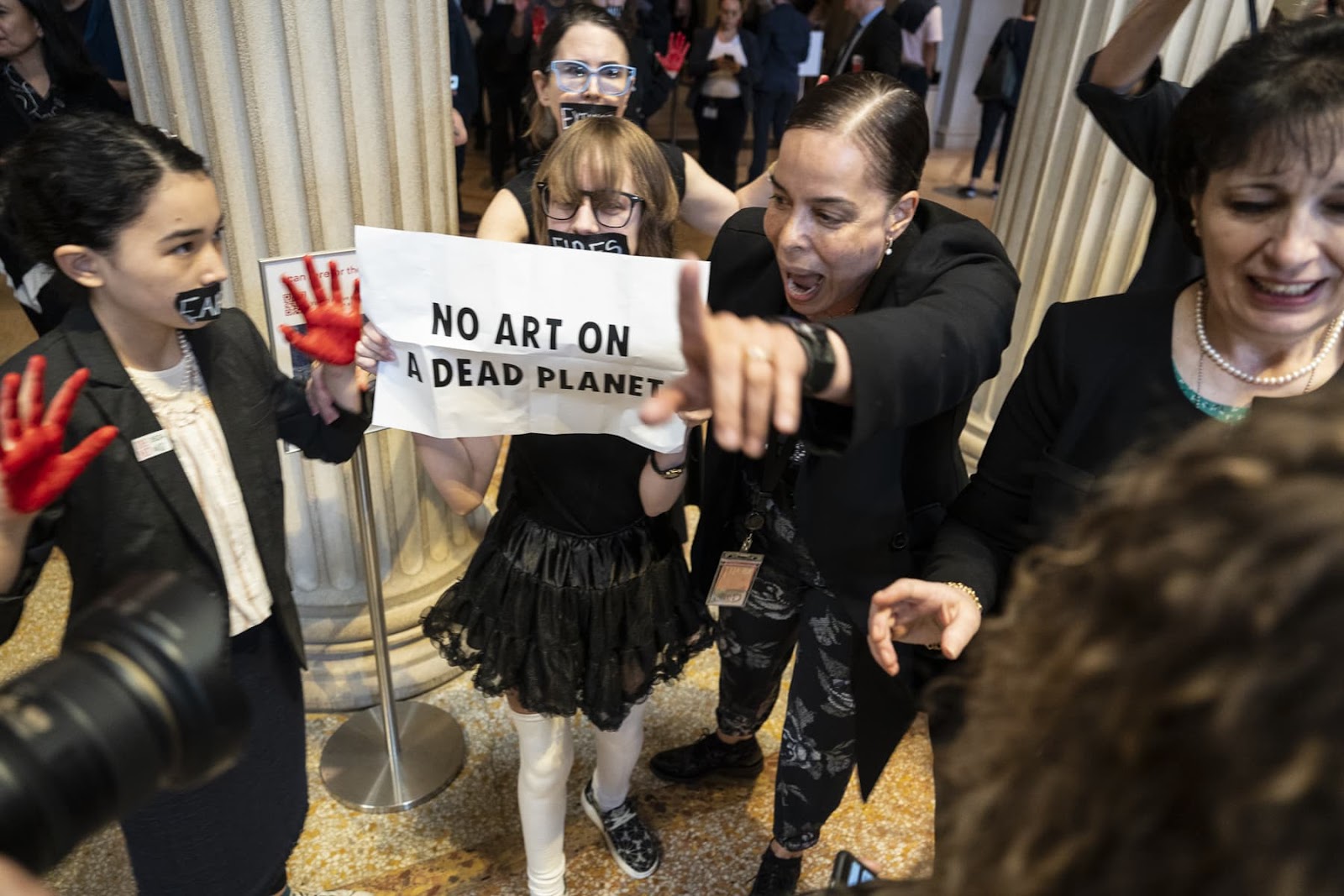 A woman screams for the acitvists to leave but they remain, tape over their mouths, one with a banner saying no art on a dead planet