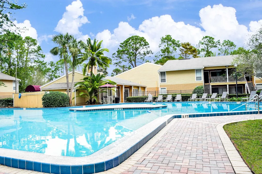 Apartment complex with a clear blue pool, lounging area, and palm trees, showcasing sunny weather.