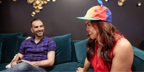 Una mujer con cabello largo está sentada en un sofá con una blusa roja y un sombrero con los colores amarillo, rojo, azul y verde de Google. Hay un hombre sentado a su lado que también sonríe.