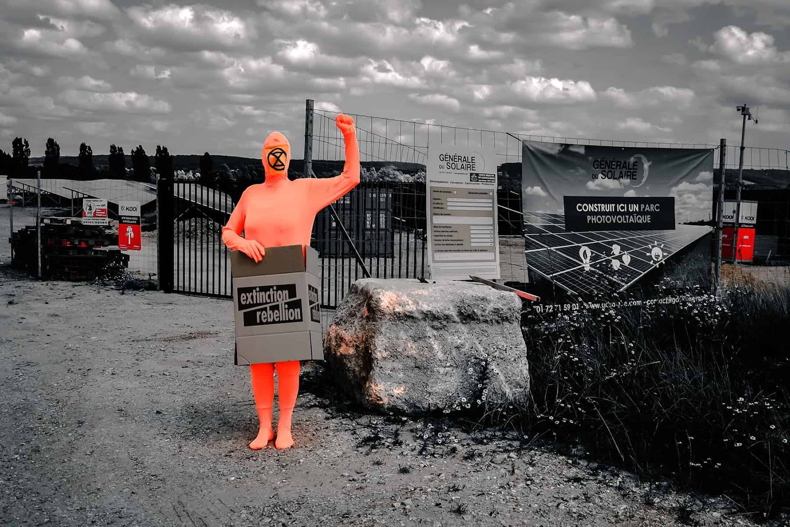 A rebel in orange body suit stands in a cardboard box before a monochrome industrial landscape