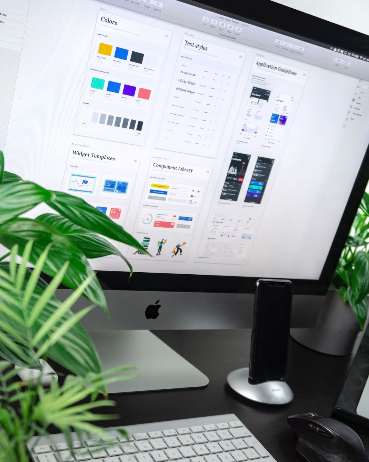 An iMac, iPhone, keyboard, and a mouse on a table surrounded with plants