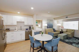 Full view of kitchen with white cabinets, round dining table with four chairs, furnished living room with tan couch and chair.