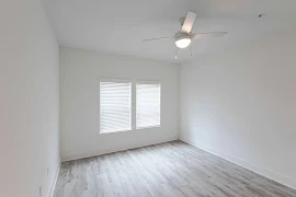 Bedroom with white walls & grey wood flooring. Two large windows are on one wall with a white ceiling fan with domed lighting