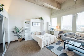 Furnished studio floorplan with three curved windows, a neutral color scheme, and tall ceilings with exposed wood