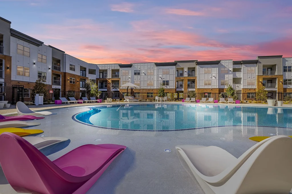 Resort-style swimming pool at dusk with blue umbrellas with lights and picnic tables, and BBQ grills