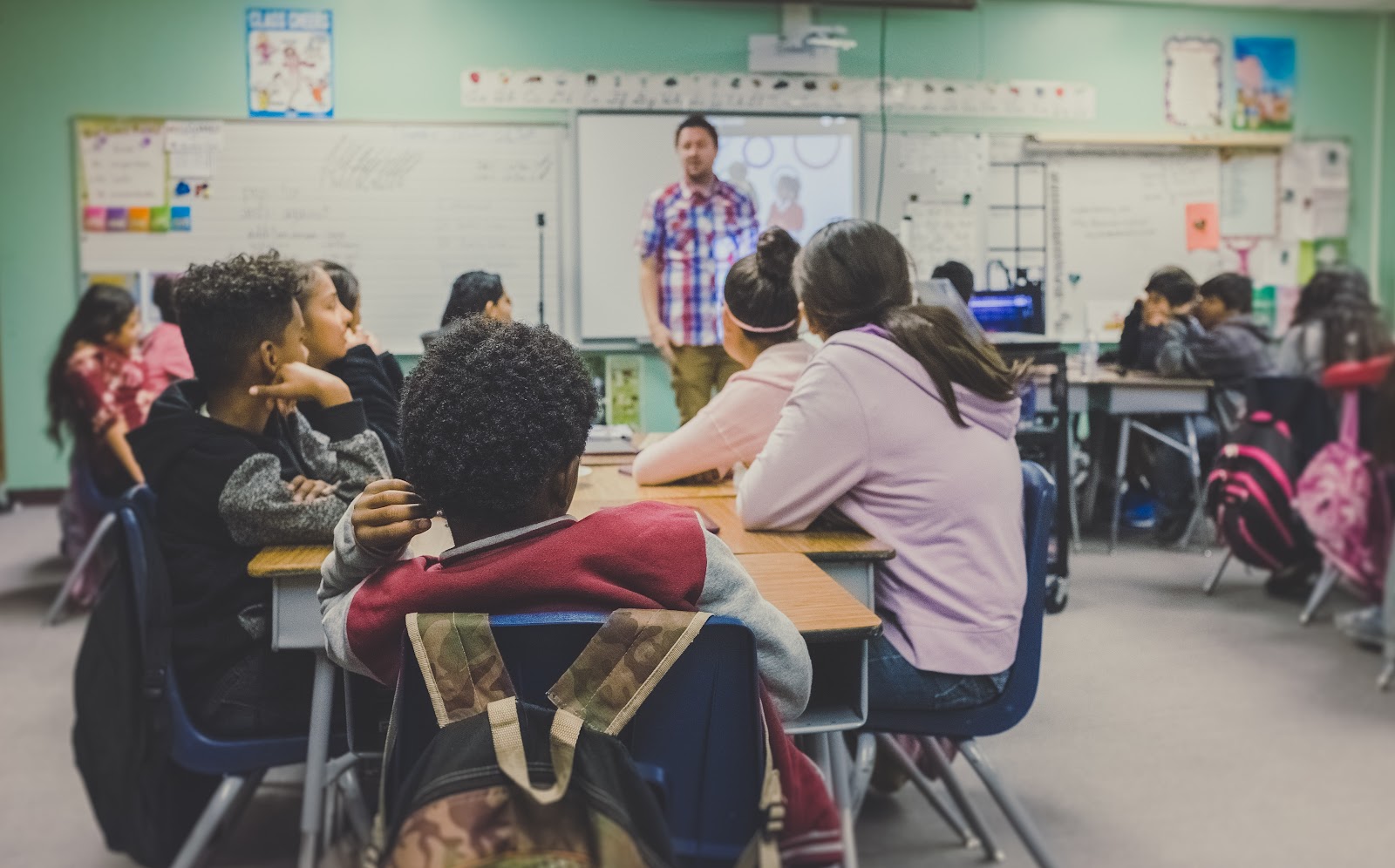 A classroom of middle-school-aged children listens as someone addresses them from in front of the whiteboard.  Students are seated in desks that are pushed together to form groups.