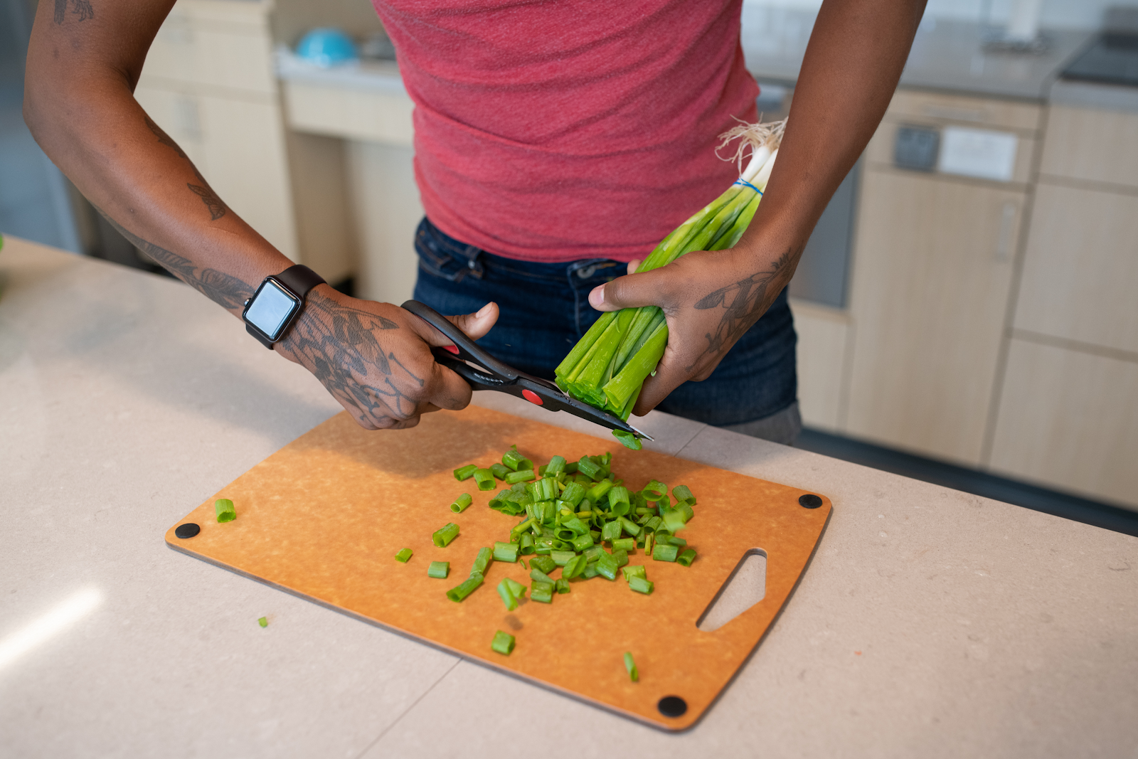 Close-up of a disabled and Black non-binary person with tattoos cutting green onions with kitchen scissors.