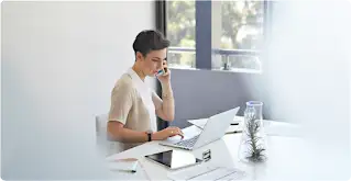 Eine Frau mit kurzen Haaren sitzt am Schreibtisch. Sie spricht in ihr Smartphone und hat einen Laptop sowie ein Tablet vor sich.
