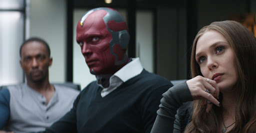 Vision wearing normal clothing. A V-neck sweater over a collared shirt. Scarlet Witch is in front of him and Flacon is behind him