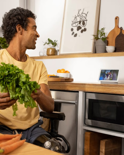 A man in a wheelchair cooks a meal on a stove while interacting with friends on a Nest Hub on the kitchen counter.