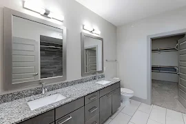 Modern apartment bathroom with dual sinks, granite countertops, and a view into a walk-in closet with built-in shelves.