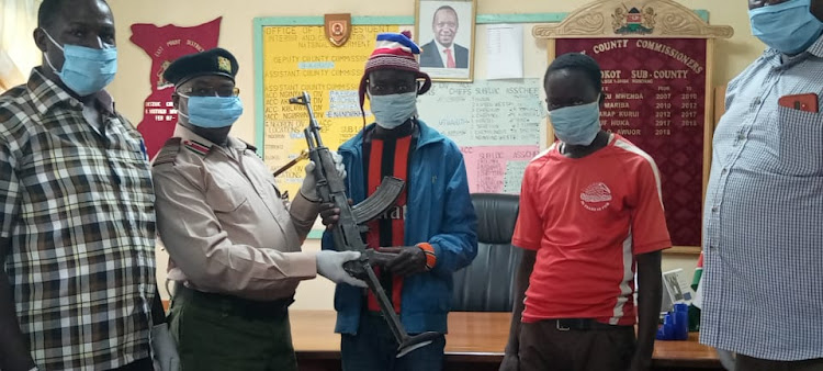 East Pokot Deputy County Commissioner Benson Karani (second left) receiving the AK-47 gun without bullets from a reformed bandit at Chemoling’ot on Saturday.