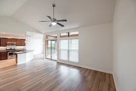 Apartment living room with wood-inspired flooring, patio doors, window with blinds, and a ceiling fan overhead 