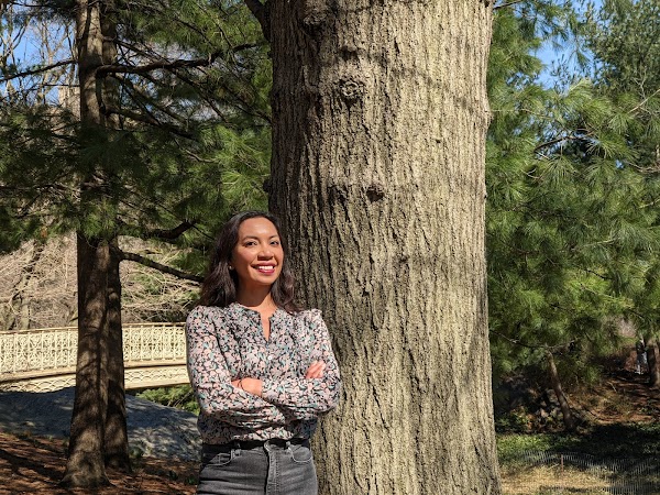 Google Research 项目经理 Ruth Alcantara 在树前交叉双臂面带微笑，身着长袖上衣和牛仔裤。