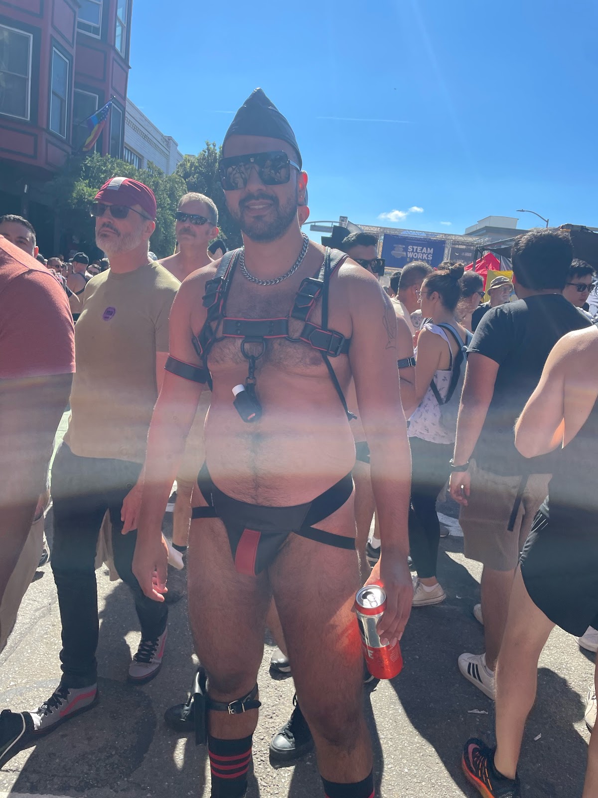 Phil DuChamp wearing a black and red leather harness and jockstrap posing for a photo at Folsom Street Fair 2023 in San Francisco