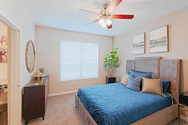 Model bedroom with light beige walls and carpet, window with blinds, and a ceiling fan