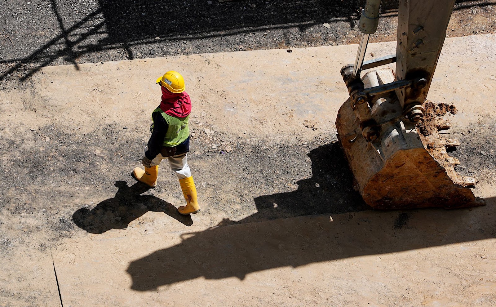 descrição: na foto observamos um homem em uma obra, ele está vestindo equipamento de segurança e se encontra ao lado de uma pá de algum maquinário que não é possível ver na imagem