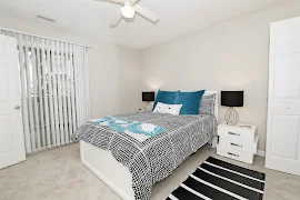 Model bedroom with black and blue comforter, plush carpet, sliding glass door with blinds, and white side tables