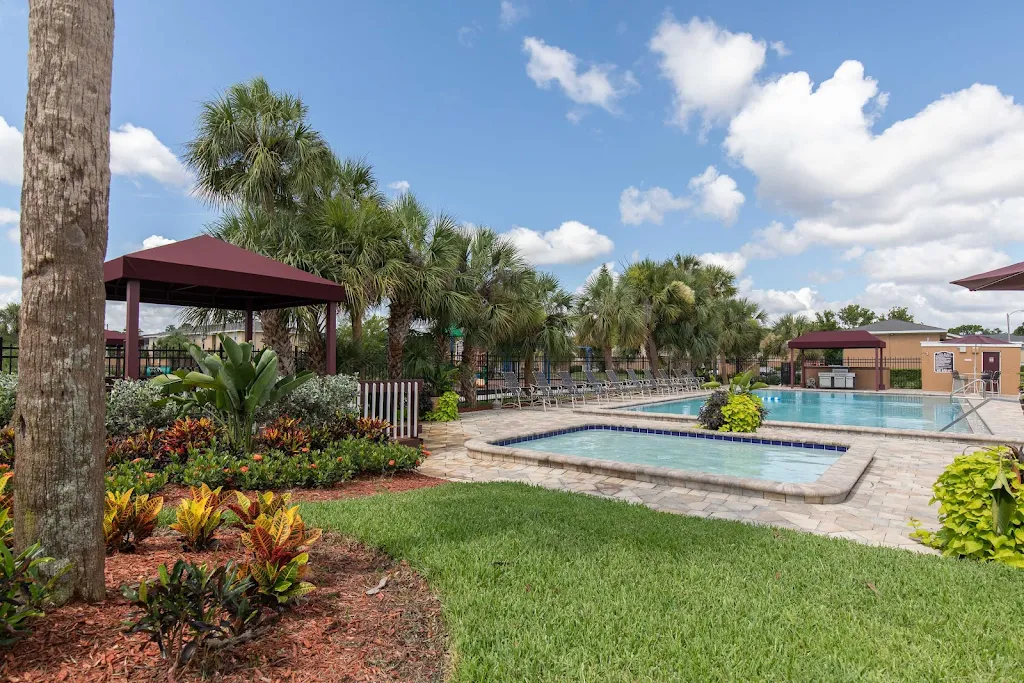 Lush apartment complex with a pool, hot tub, loungers, and tropical landscaping under a blue sky.