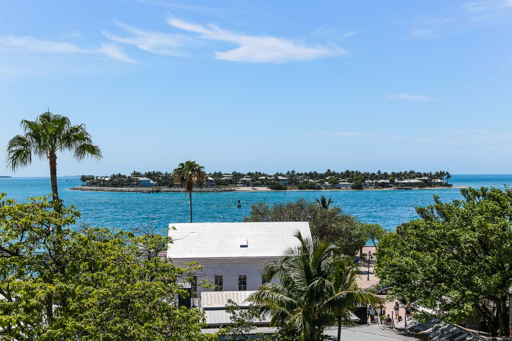 A key is seen off the coast surrounded by teal sparkling water typical of the Florida Keys. There is lots of greenery everywhere and some buildings are visible on the key.