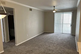 Empty apartment interior with gray carpet, white walls, ceiling fan, and sliding glass door leading to balcony.