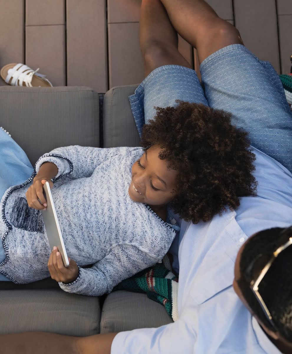 Una bambina guarda lo schermo di un tablet con la testa appoggiata sulle gambe del papà mentre sono all'aperto.