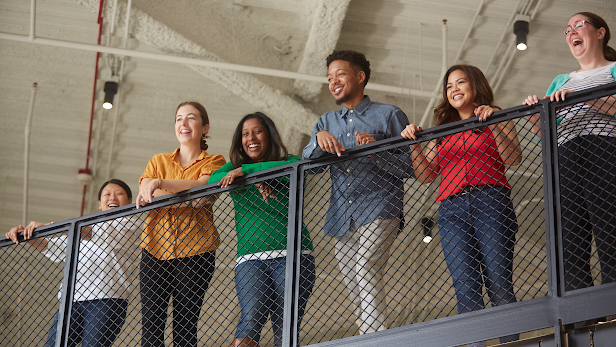 Un grupo de empleados de Google felices con camisas de colores brillantes miran hacia abajo desde la barandilla de la escalera de un lugar de trabajo