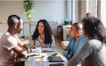 Quatro membros de uma equipa sentados à mesa a ter uma reunião de trabalho e a tomar café com notebooks, tablets e telemóveis sobre a mesa.