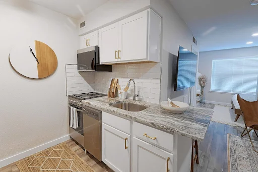 Model kitchen with white shaker cabinets, brass pulls, wood floor, stone counter, stainless steel appliances, tile backsplash