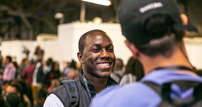 A man smiling in a crowd wearing a backpack and vest