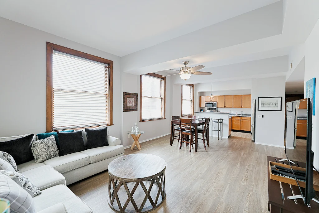 Model unit living room, facing the kitchen, with wood plank floors, white walls, windows with wood trim, and furniture