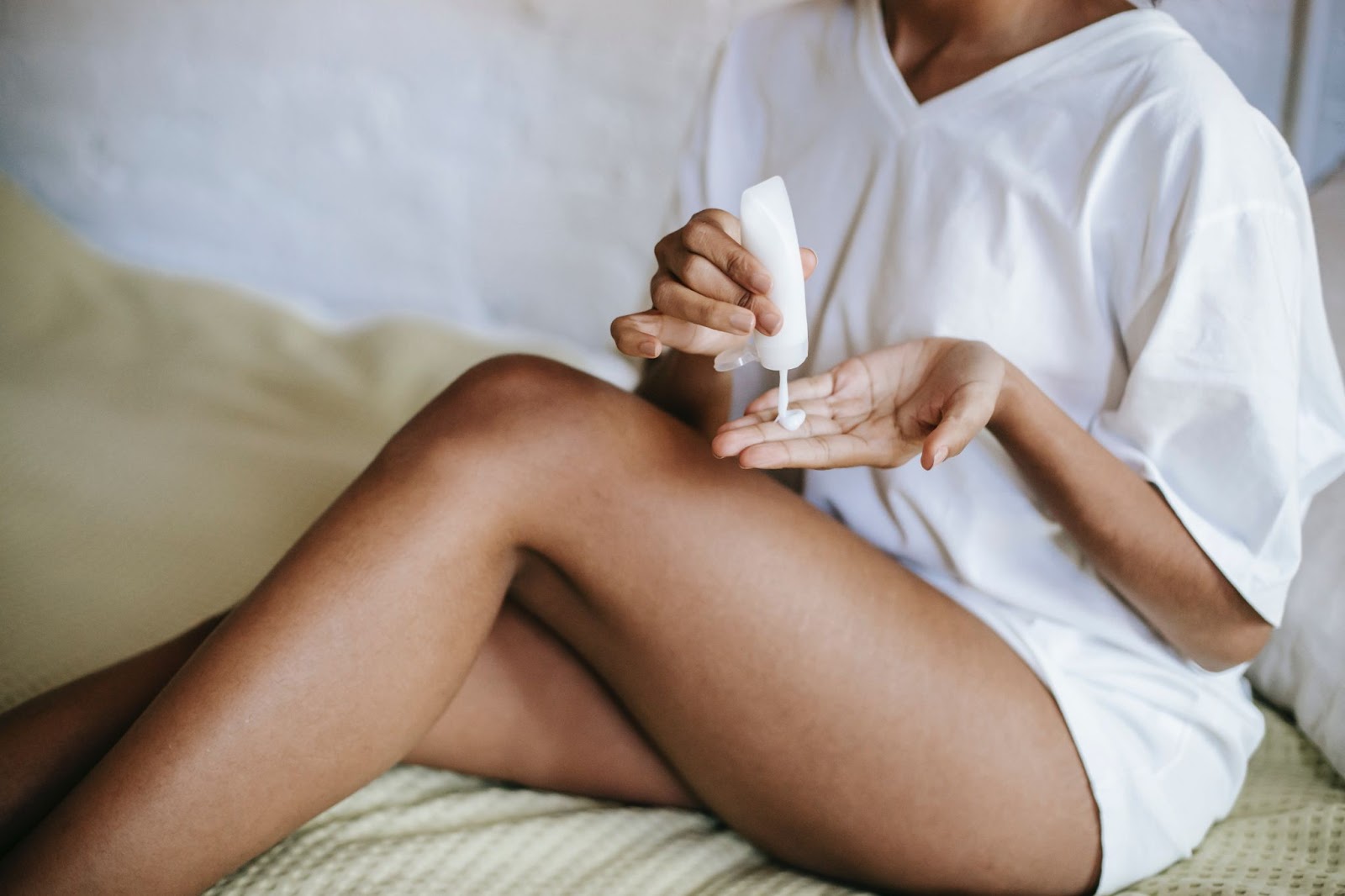 a imagem mostra uma mulher negra, em cima de uma cama, com uma camiseta larga branca e um hidratante corporal na mão esquerda. 