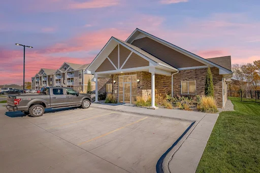 Clubhouse exterior with tan siding, brick detailing, and foliage next to a parking lot 