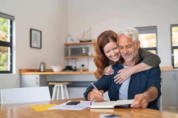 Loving woman embracing senior man Loving mature wife embracing husband from behind while writing in book. Happy middle aged couple making to do list of purchases and discussing future plans. Cheerful senior man working at home on wooden table with beautiful woman hugging him from behind, copy space. Elderly making lists for moving stock pictures, royalty-free photos & images