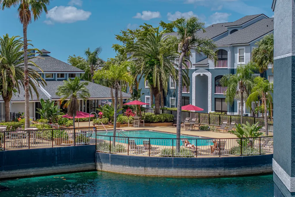 Luxurious apartment complex with pool, palm trees, and loungers by a serene water body, under a clear blue sky.