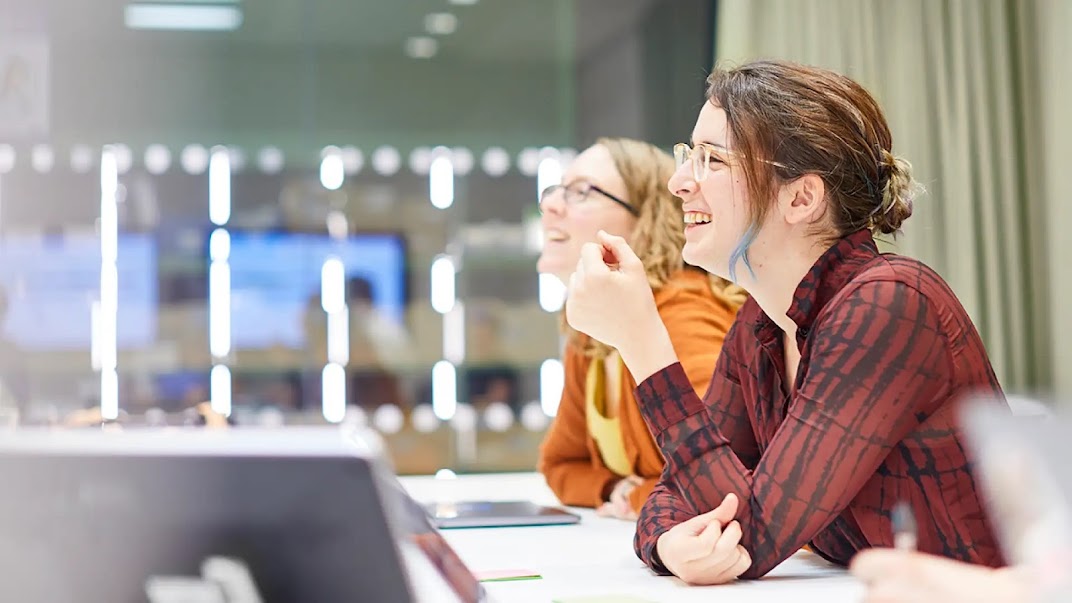 Two women in a meeting room, viewed from the side, and smiling at something we can't see. There are laptops on the table and the reflection of a presentation in the window behind them.