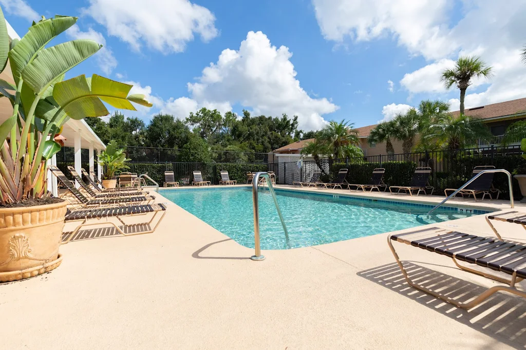 Apartment complex pool with lounge chairs, surrounded by lush greenery under a sunny sky.