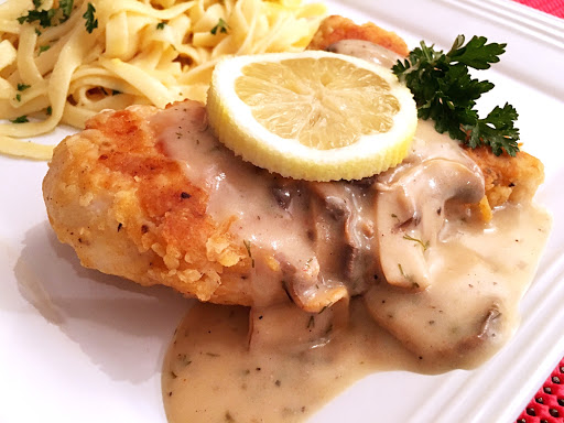 Breaded chicken breast topped with sauce, a slice of lemon and garnish with parsley on a white plate with pasta in the background. 