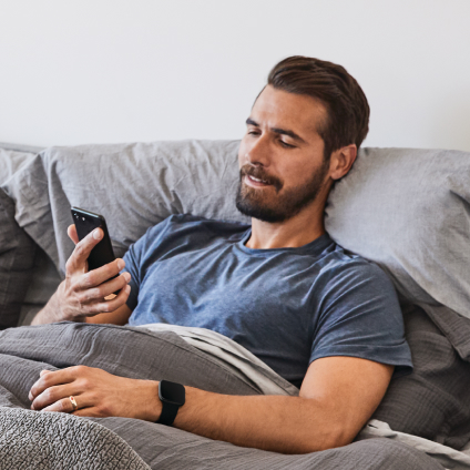 Un homme allongé dans son lit avec une montre connectée Fitbit au poignet et qui regarde son téléphone