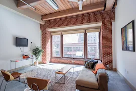 Living room with three large windows, brick accent wall, couch, mounted TV, coffee table, and additional decor