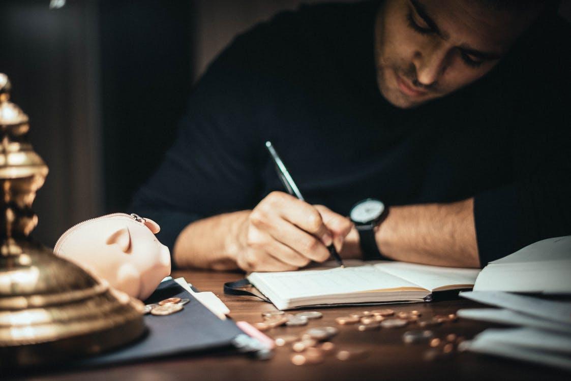 Free Crop elegant man taking notes in journal while working at desk with coins and piggybank in lamplight Stock Photo
