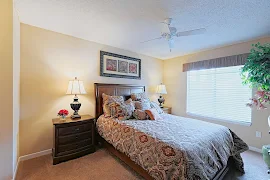 Cozy apartment bedroom with a queen bed, nightstand, lamps, artwork, and a window with blinds.