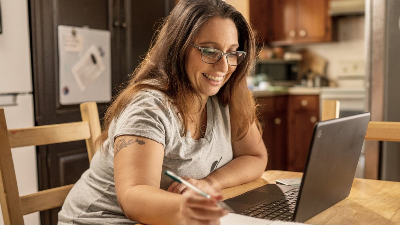 A woman sits at her desk working on a laptop. She's smiling.