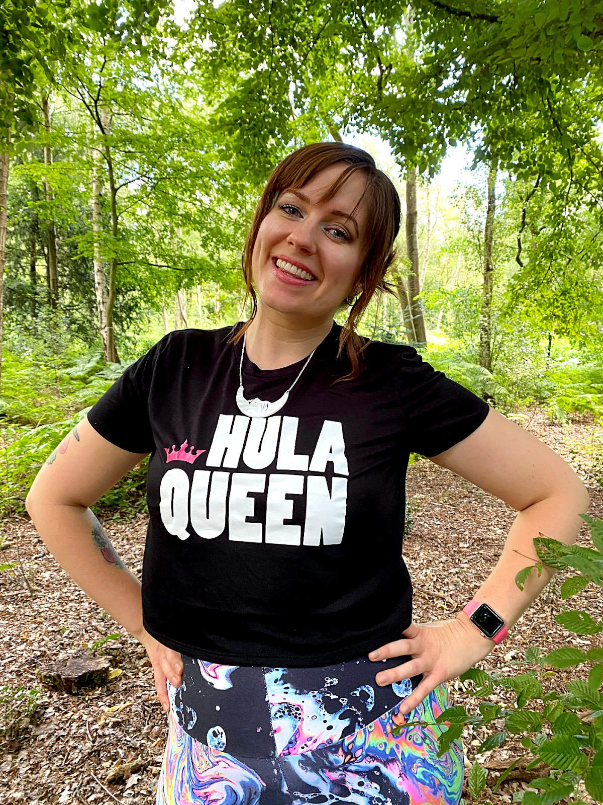 Sally, a white woman with dark hair and a fringe is wearing a black 'hula queen' top and has her hands on her hips. she is standing in a wood clearing.