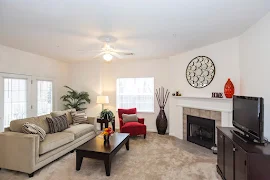 Model unit living room features neutral carpet, white walls, white ceiling fan, corner fireplace, and patio door with blinds