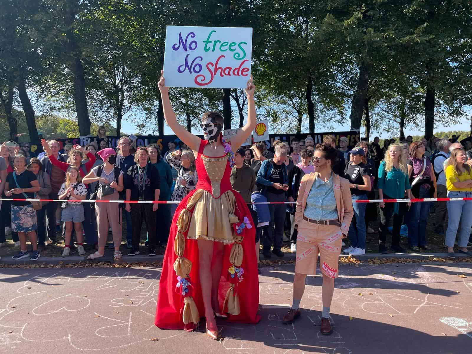 A fabulous drag queen and drag king stand during the A12 Blockade. The drag queen holds up a sign "No trees No shade".