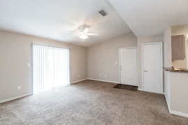 Living area with sliding glass door with vertical blinds, ceiling fan with light fixture, and neutral carpeting and walls.