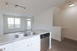 Kitchen with light cupboards, neutral counter with stainless appliances with the living room in the background.