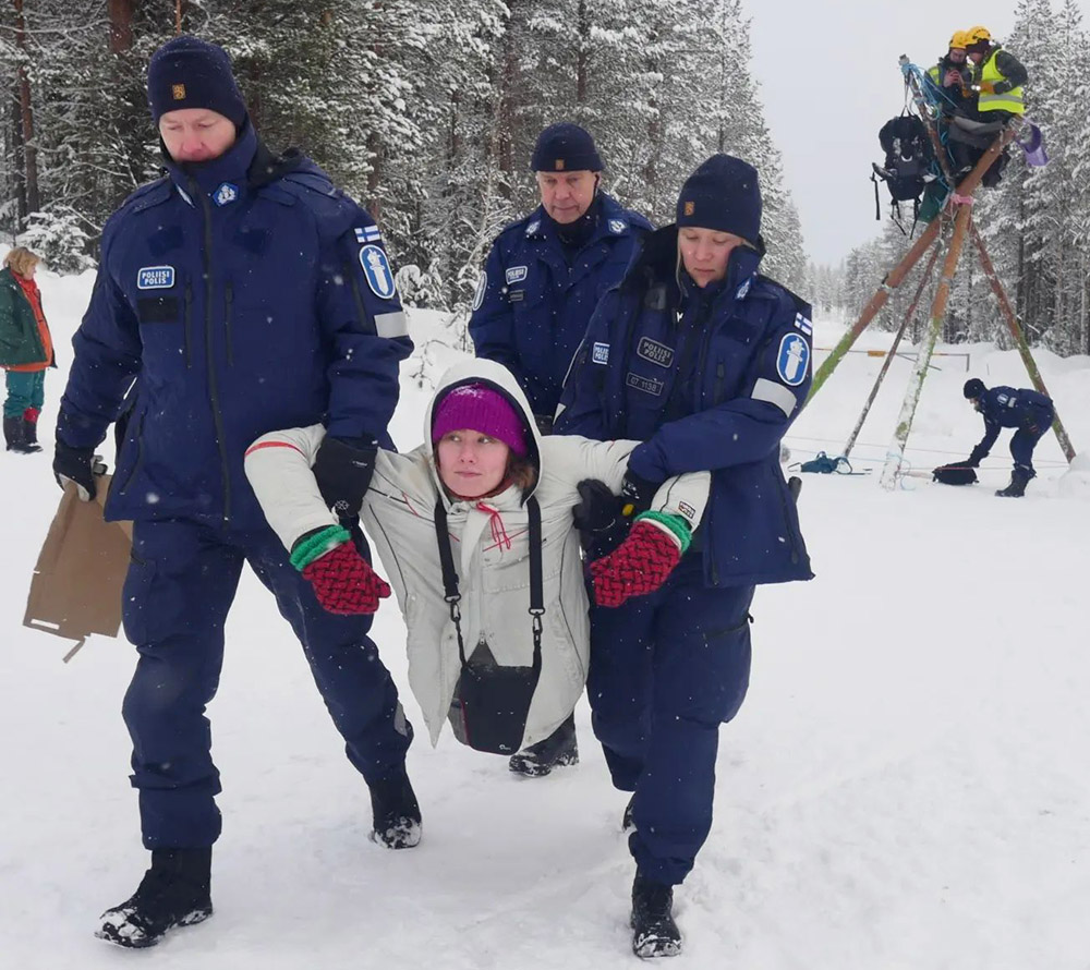 Three police haul a young woman down the snow-covered road while two more activists sit on top of a tripod behind