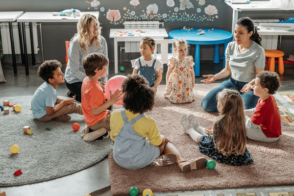 A group of preschoolers sitting on a rug with two adults.